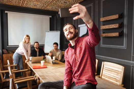 A man in an office, taking a selfie infront of his colleagues A man taking a selfie